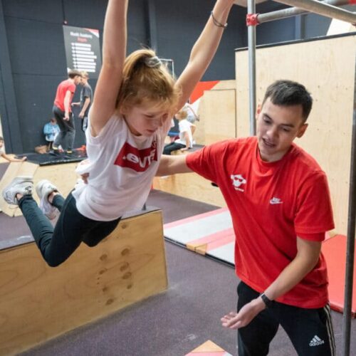 Een meisje die een zwaai oefent in de freerunhal en wordt geholpen bij een workshop freerunning