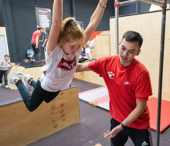 Een meisje die een zwaai oefent in de freerunhal en wordt geholpen bij een workshop freerunning